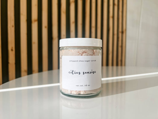Jar of whipped shea sugar scrub labeled 'citrus sunrise' on a white surface with a wooden background.