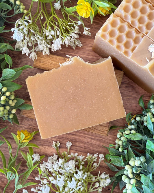 Two Beige bars of soap on a wooden surface with greenery and flowers.
