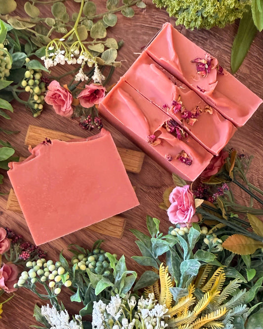 Two pink soap bars with floral elements on a wooden surface surrounded by flowers and greenery.