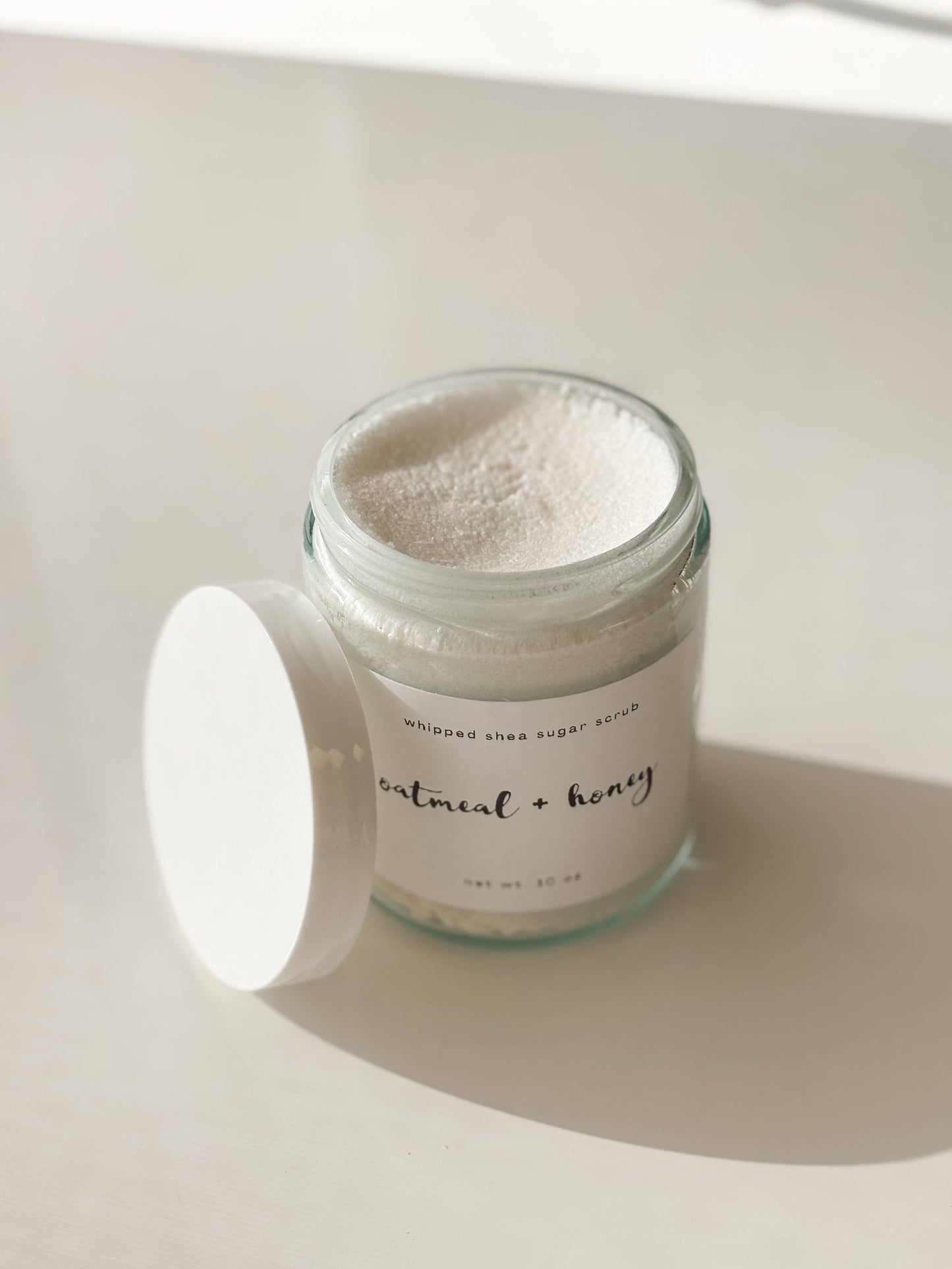 Jar of sugar scrub with a white lid on a light background. Labeled "oatmeal + honey".