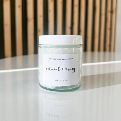 Jar of whipped shea sugar scrub labeled 'oatmeal + honey' on a reflective surface with a wooden background.
