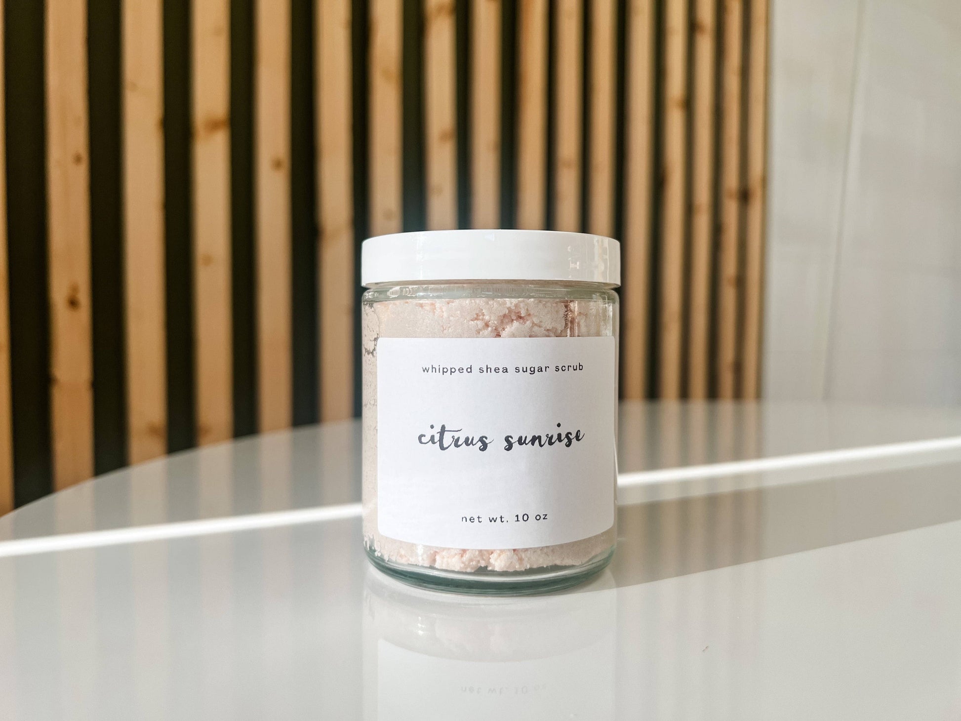 Jar of whipped shea sugar scrub labeled 'citrus sunrise' on a white surface with a wooden background.