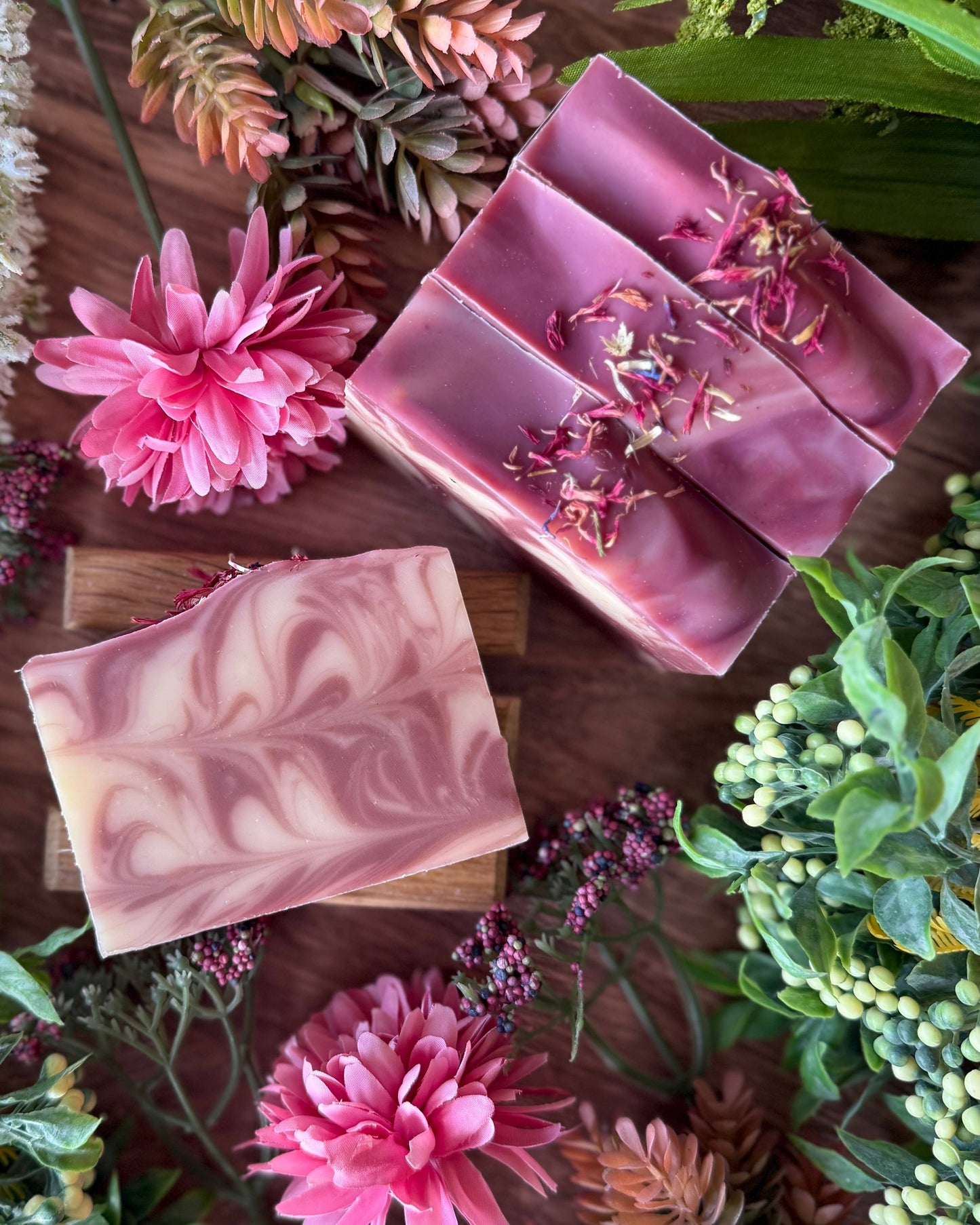 Pink soap bars with floral elements on a wooden surface surrounded by flowers and plants