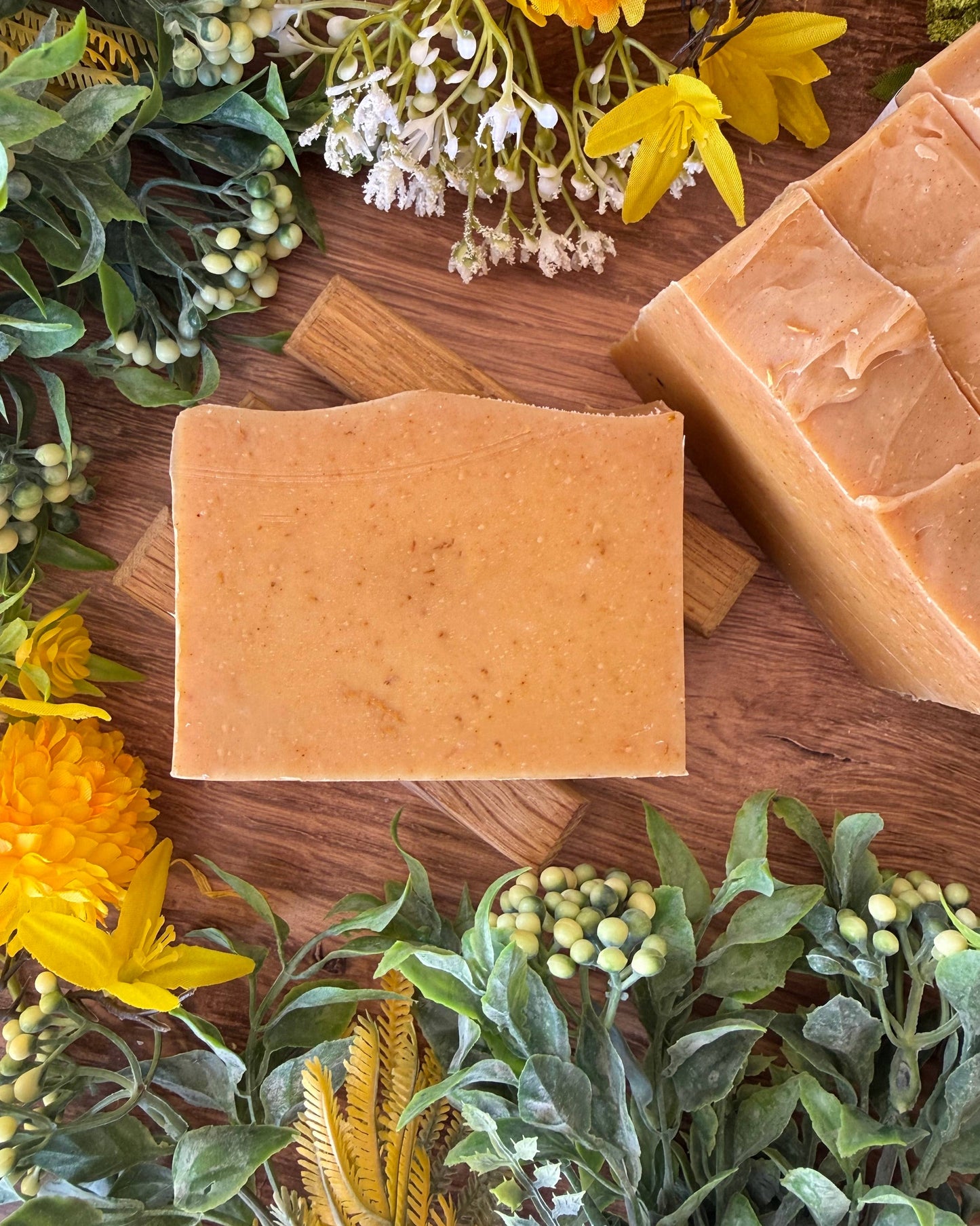 Two yellow bars of soap on a wooden surface with flowers and leaves around.