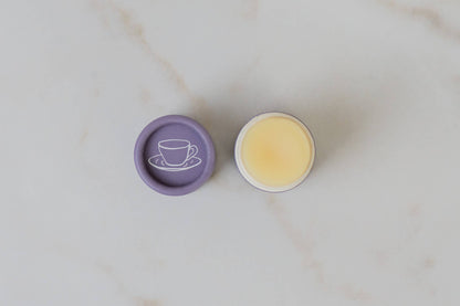 An open container of lip butter with lid laid on one side that has coffee cup logo on it. 